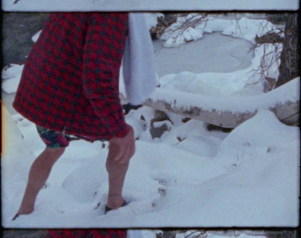 A man wearing shorts and flannel turns and waves then walks on snow covered rocks toward a stream