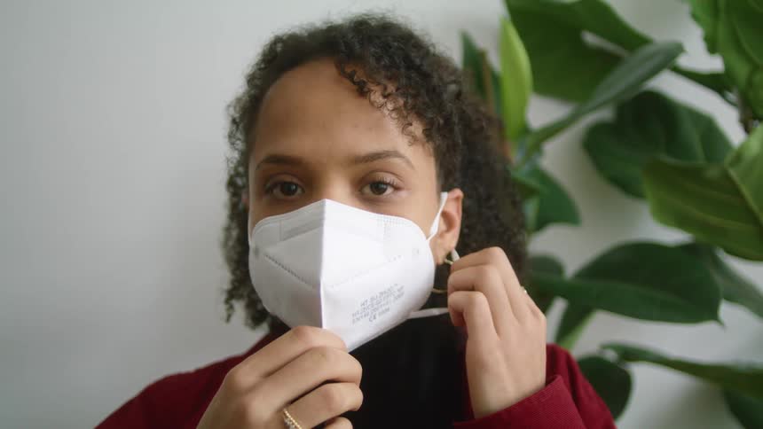 A woman putting on a white face mask in front of a potted houseplant