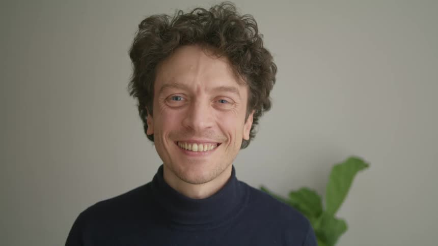 Man with navy blue turtleneck sweater takes off his white mask and smiles at camera