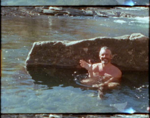 A man sits in a river leaning against a large rock as the sun shines and shimmers on the surface
