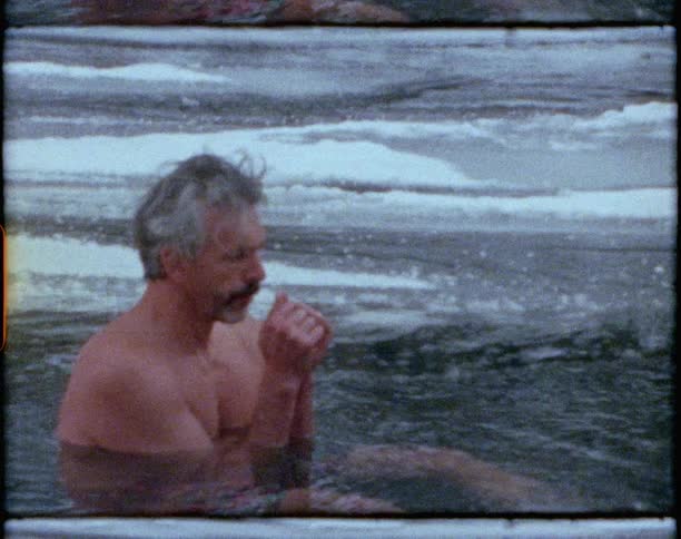 Vintage footage of a man sitting in a river in the winter blowing in his hands to warm up and running his hands filled with water through his hair