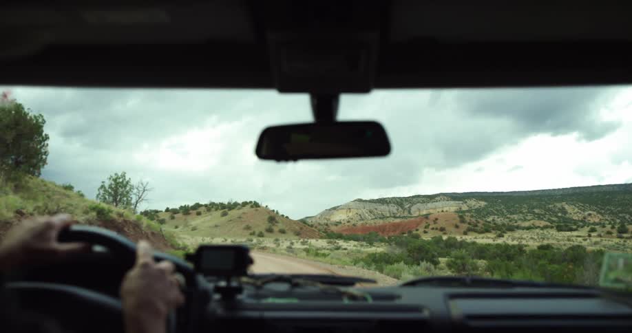 Person drives car along winding dirt road through scrub dotted hills following another car