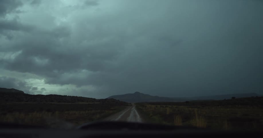 A vehicle with windshield wipers driving on a road towards a mountain range with stormy sky