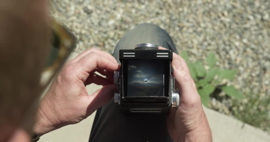 A man sits with his camera on his leg and adjusts knobs on a bright sunny day