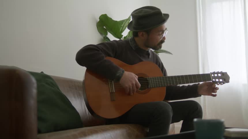A man sitting on a leather sofa in the living room of a house tunes his guitar
