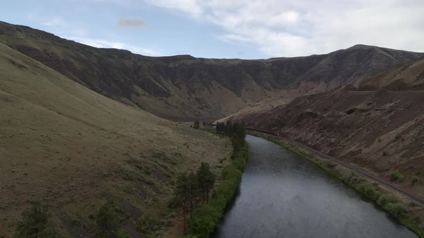A freight train slowly rounds the bend of a river valley surrounded by rolling hills
