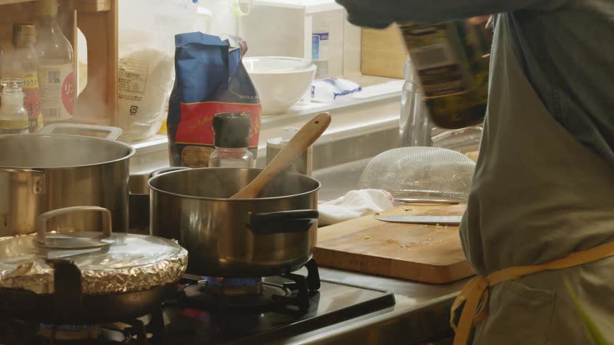 A man wearing an apron pours olive oil inside a pot boiling over the kitchen at home