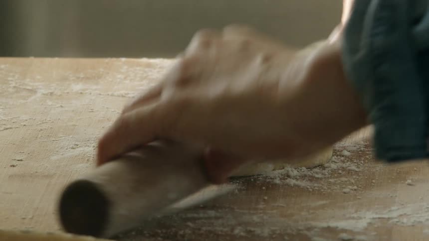 Close up of a person using a rolling pin to flatten dough over a floured wooden board