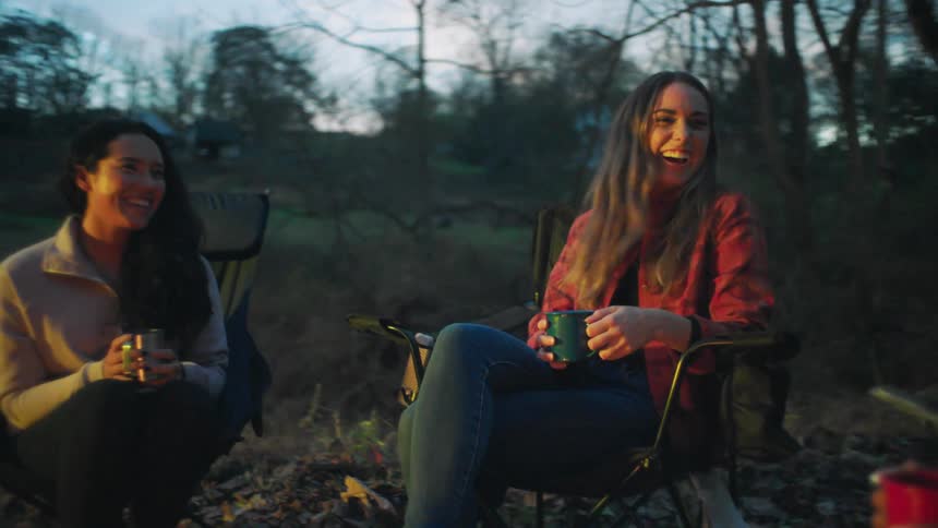 Three women sitting on camp chairs next to a fire with trees the sky and clouds in the background