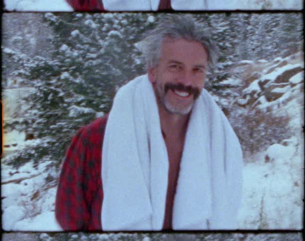 Man wearing shorts and open shirt hikes through snow away from river in forest
