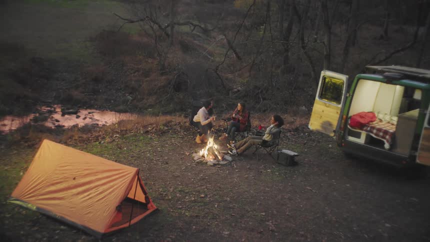 Three women gather around a campfire and make a toast with coffee mugs