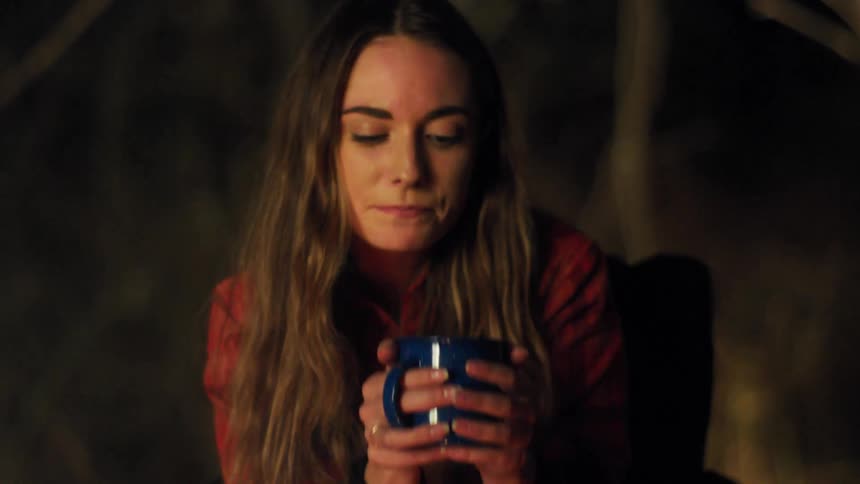 A woman sitting on a chair and drinking from a mug with smoke in the foreground and trees in the background
