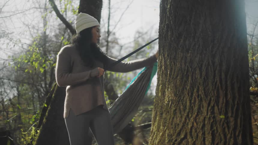 A woman tying a string of a hammock to a tree in a forest at daytime