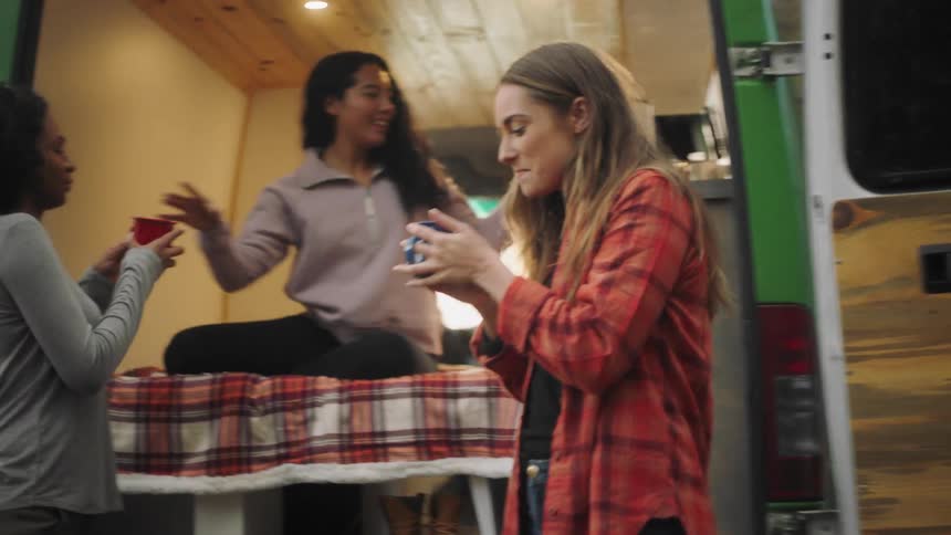 A woman sitting in the back of a van hands out drinks to a pair of women before taking a drink of her own and exiting the van