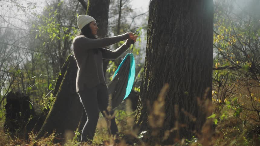 A young woman hangs a hammock in between two trees in a forest