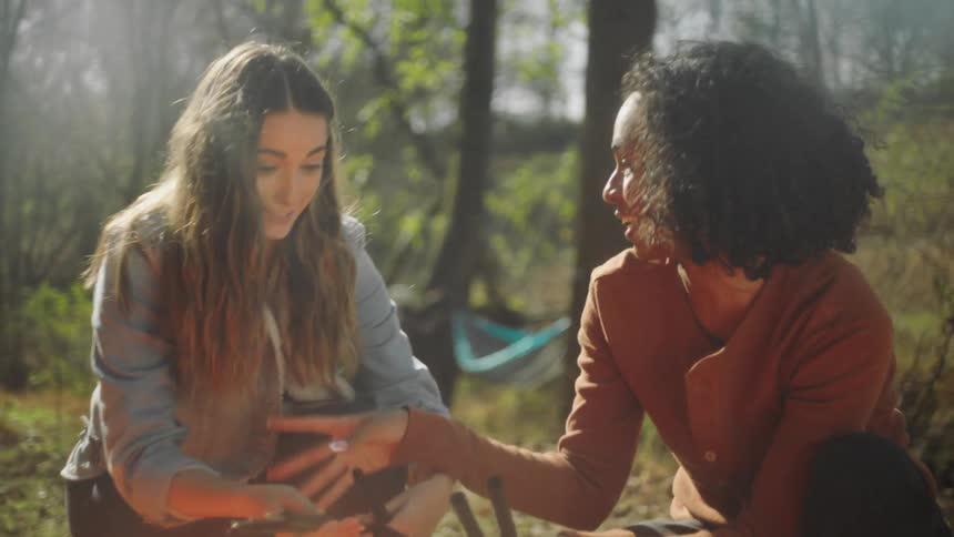 Two women pitching a tent with a woman behind them next to a hammock with trees the sky and clouds in the background