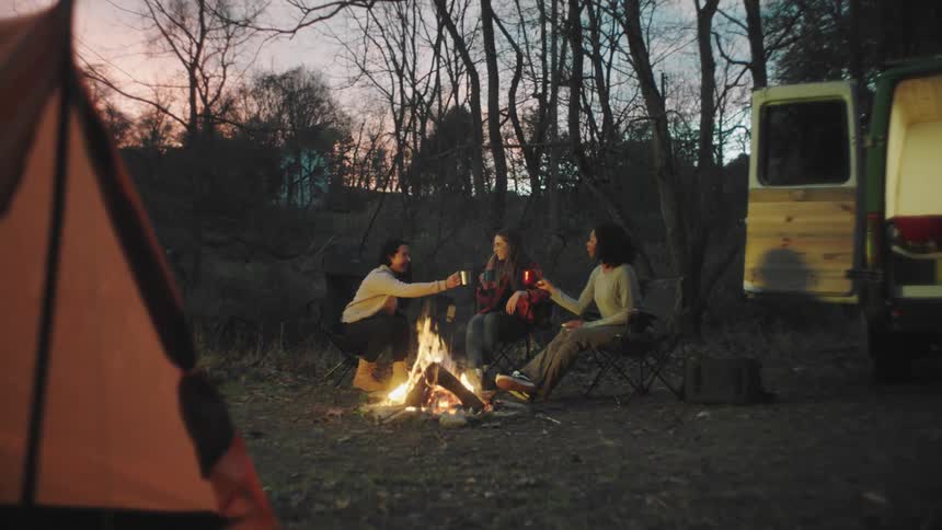 Three friends sit around a camp fire and clink mugs as they toast