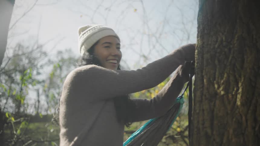 A woman smiling and shaking her head while hanging a hammock from a tree on a sunny day