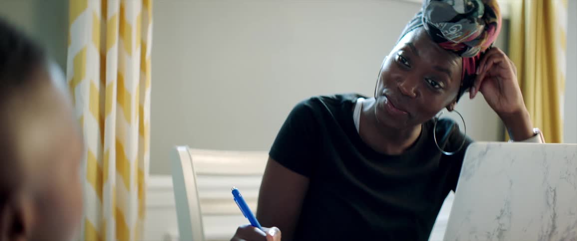 A smiling woman sitting before a laptop at a table speaks with a child sitting across from her