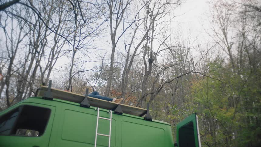 A person lies on the roof of a green camper looking up at the trees