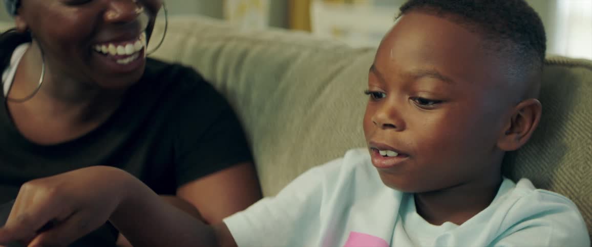 A mother smiling sitting with her son on a couch while he talks and points in front of him