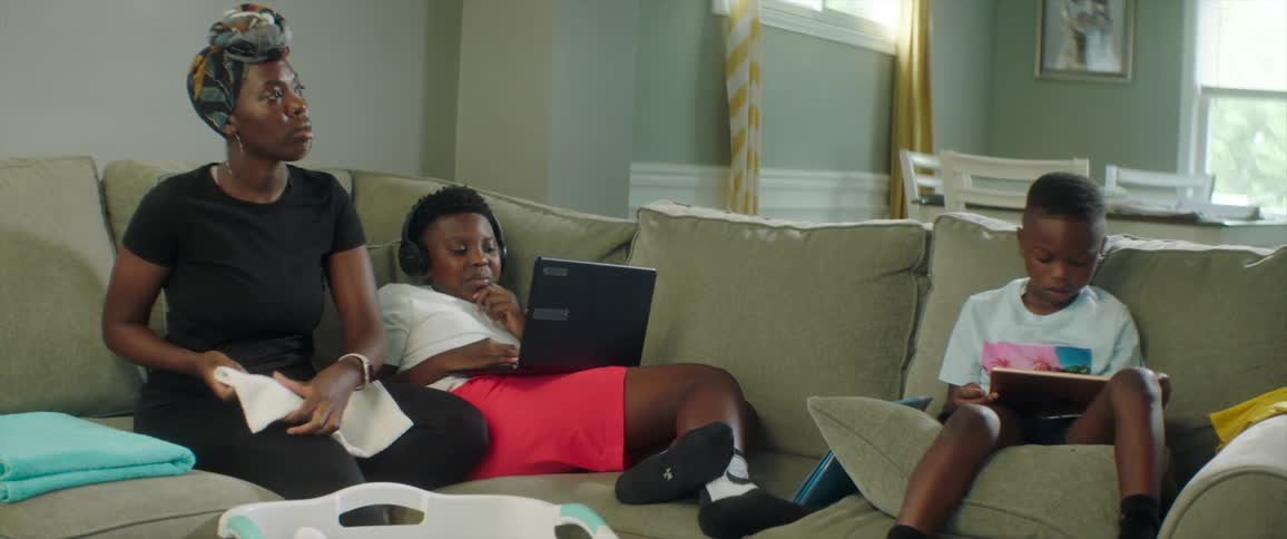 Children sit on couch viewing device while mother folds clothing