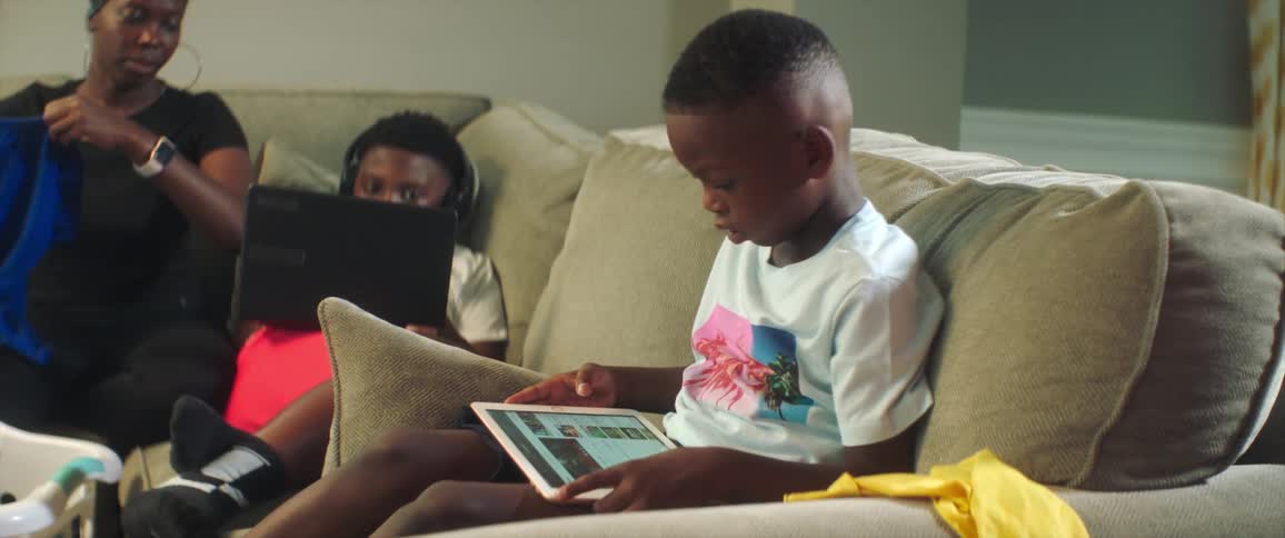 A young boy sitting on a couch looking down at a tablet next to his mother folding laundry and his brother on a laptop