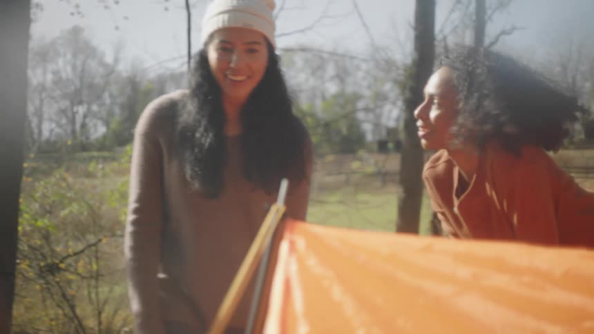 A young woman approaches a pair of women standing around a makeshift tent in a woodland during the day