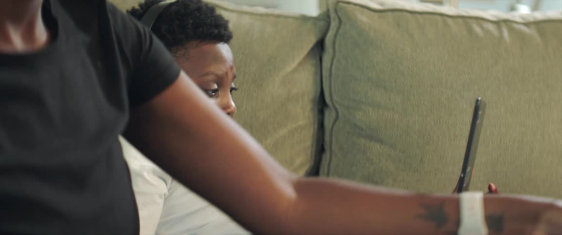A boy wearing headphones watches the screen of a laptop while sitting on a sofa next to a woman folding laundry
