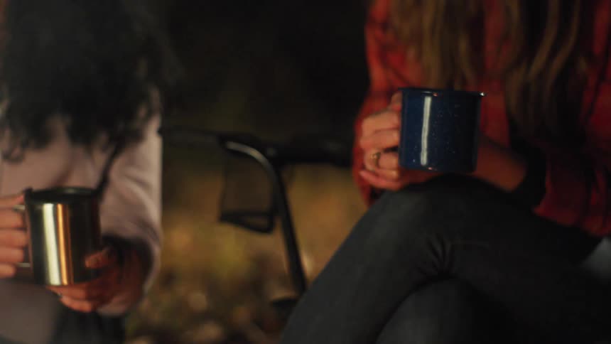 Two women hold coffee cups while sitting outside around a fire at nighttime