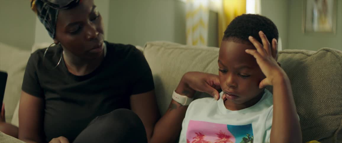 A woman sitting on a sofa next to a boy taps the screen of his tablet before adjusting his shirt
