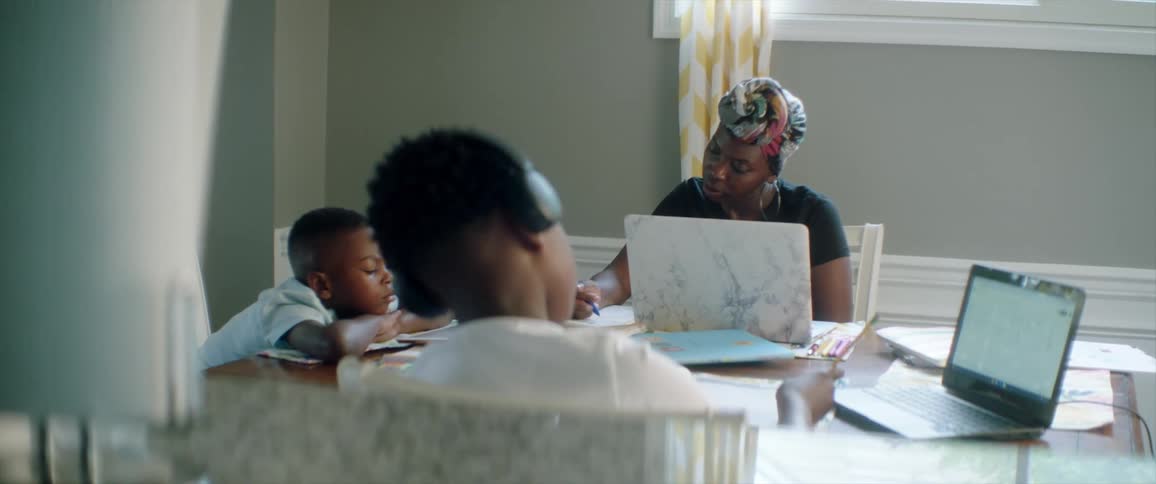 A woman uses a laptop as children write at the table
