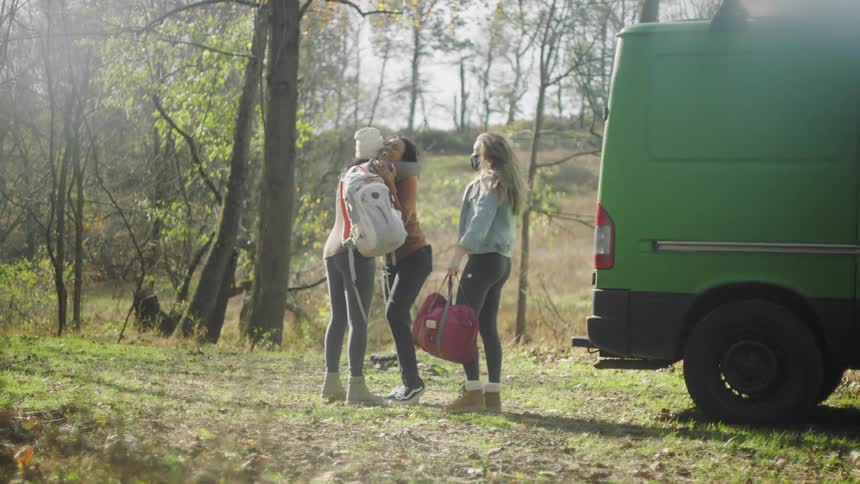Three woman next to a vehicle hugging with trees the sky and clouds in the background