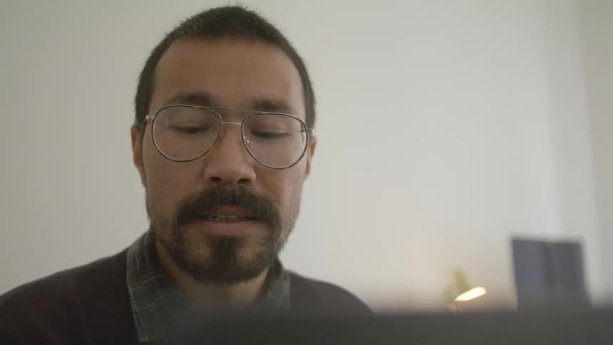 A man sitting down indoors reaches over to a cellphone and makes a phone call
