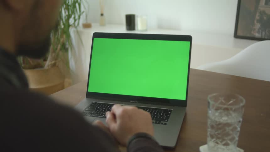 A man sitting at a table in a house gestures while participating in a video conference using a laptop