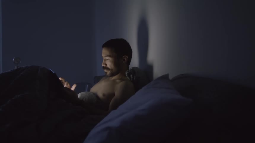 Shirtless man with beard lies in bed and uses tablet in dark room