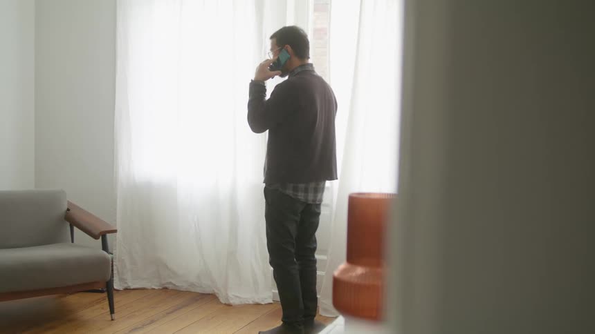 A man putting on eyeglasses and pacing while talking on a phone in front of a window