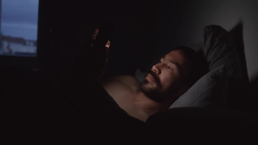 Shirtless man with beard lies in bed and uses smartphone in the dark bedroom