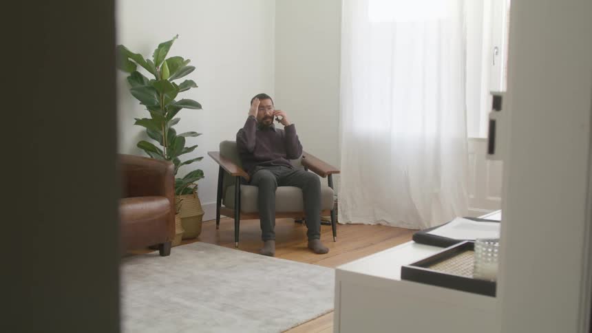 A man in a room talking on a smart phone with a wall and windows in the background