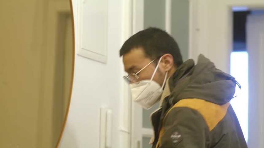 A man at home stands in front of a mirror as he puts a mask on and then walks out the door