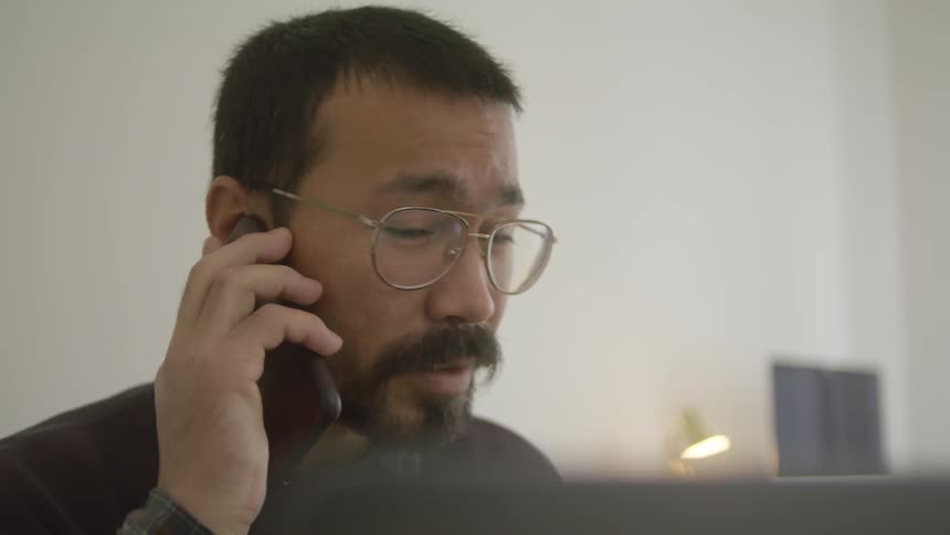 Bearded man wearing glasses ends a call on his smartphone and continues working
