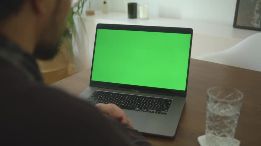 A man at home sits and looks at a green screen on his laptop