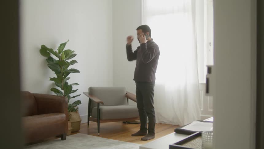 A man speaks on a smartphone while pacing in front of a window in the living room of a house before tossing the phone onto a chair