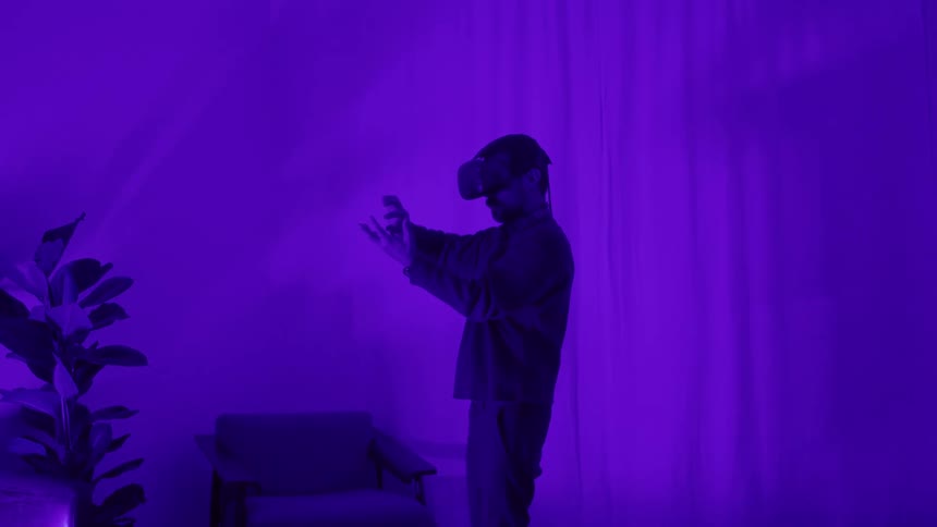A man wearing virtual reality goggles manipulates objects with his hands while standing in a room