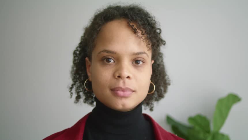A woman with a turtleneck sweater standing in a room with a wall and a plant in the background