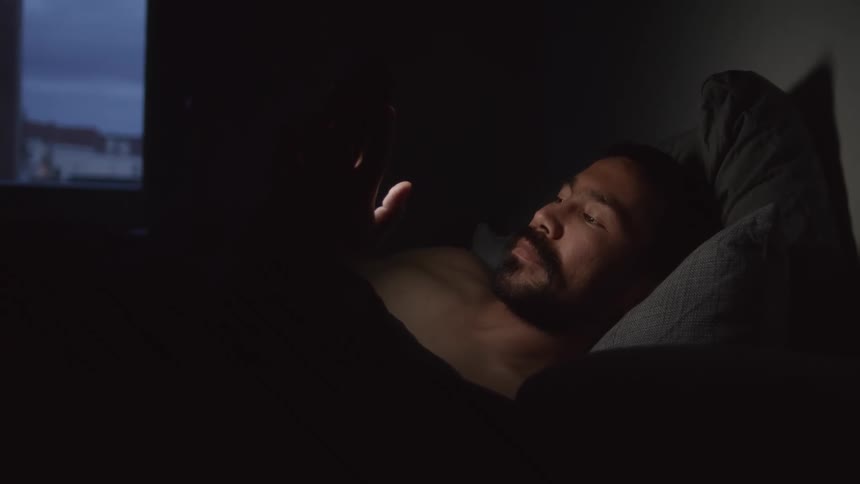 A man lying in bed in a dark room taps the screen of his smartphone while laying in bed before the screen goes dark