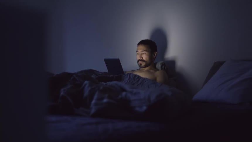 A man in a bedroom sitting in bed with a blanket and pillows looking at a tablet