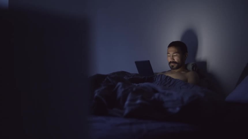 A shirtless man reading in bed in a dark bedroom