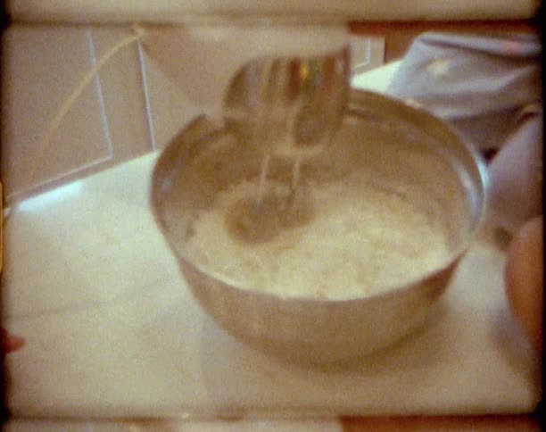 Little girl sitting on the kitchen counter top helps her mother mixing dough using a hand mixer