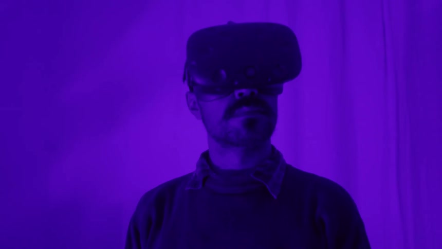 A man with a vr headset in a room with purple light with a wall and a plant in the background